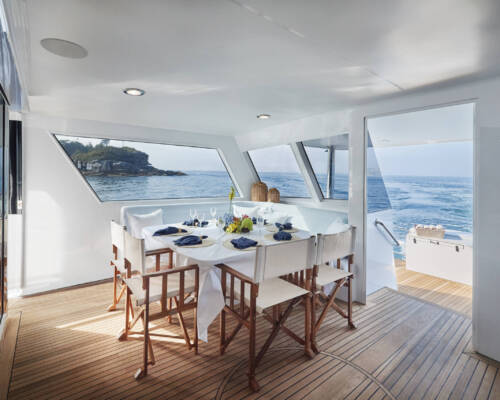 ShoreThing_Main_Deck_Dining A set dining table with white chairs inside a modern yacht overlooking the ocean